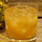 A glass of pineapple juice with ice next to a wooden spoon.